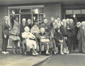 a group of patients and staff outside a Home for the Elderly. Emma's grandmother is waving to the camera while other wave or smile.