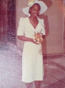 A bride stands in a knee-length white dress with a 70's style wide brimmed hat, clutching a posy of flowers